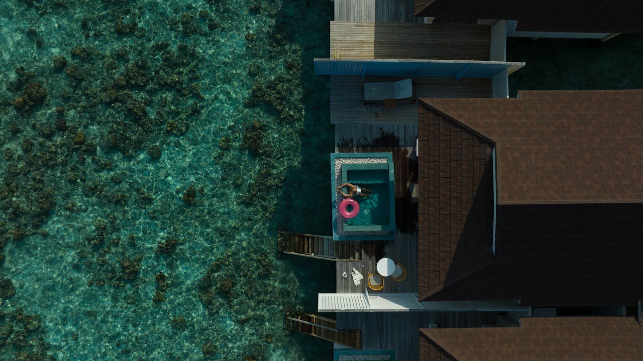 Stunning aerial view of a Maldives overwater villa with clear lagoon, featuring a bright pool float.